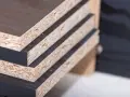 Stos płyt meblowych z widocznymi warstwami i ciemnym laminatem. Pozwala to ocenić ich wymiary i grubość.