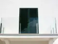 Nowoczesny balkon z przeszkloną balustradą i ciemnozielonymi okiennicami. Okapnik chroni przed deszczem.