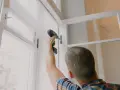 Mężczyzna wkręca wkręt w ramę okna za pomocą wiertarki.