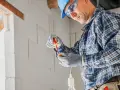 Elektryk w kasku i okularach montuje oprawkę żarówki. Zastanawia się, ile za punkt elektryczny.