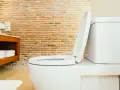 Biała miska WC z podniesioną deską, widoczna w łazience z ceglaną ścianą. Określenie jaka wysokość miski WC od podłogi jest kluczowa dla wygody.