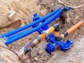 Instalacja gazowa w wykopie. Widoczne niebieskie rury osłonowe, pomarańczowe rury gazowe i niebieskie zawory. Koszt instalacji gazowej zależy od wielu czynników.