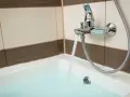 Chromowana bateria wannowa z wylewką i słuchawką prysznicową, z której leje się woda do wanny. Widać fragment kafelków i wannę z wodą.