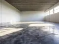 Pusta hala z betonową podłogą, która lśni jak polerowana żywica. Światło wpada przez wysokie okna, tworząc na niej refleksy.
