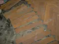 Drewniany parkiet w jodełkę, częściowo podniesiony, odsłaniający podłoże.