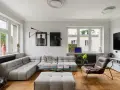 Nowoczesny salon z szarym narożnikiem, szklanym stolikiem i brązowym fotelem. Widać dzieła sztuki, rośliny i telewizor. To przykład udanego **retrofit** mieszkania.