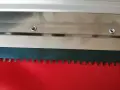 Metalowa rakla z ząbkowanym ostrzem, gotowa do pracy na czerwonym materiale.