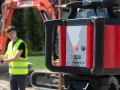 Niwelator laserowy Pulsar H na budowie. W tle pracownik w kamizelce odblaskowej i koparka.
