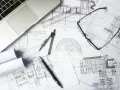 Na stole leżą plany architektoniczne, laptop, okulary i przybory kreślarskie. To miejsce pracy nad kosztorysem inwestorskim.