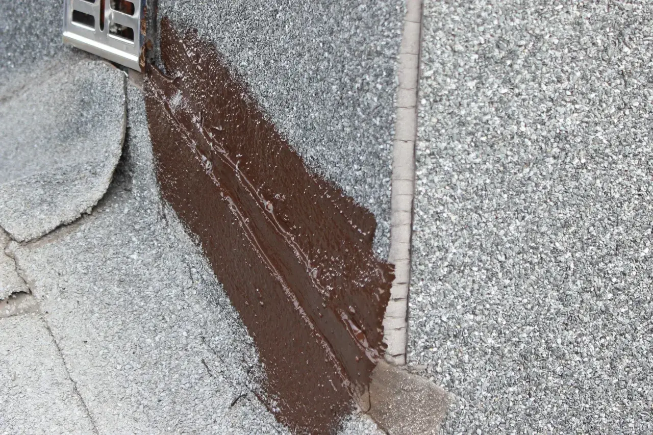 Fragment dachu pokryty papą z posypką, gdzie świeżo nałożona brązowa masa bitumiczna uszczelnia połączenie z metalowym elementem.