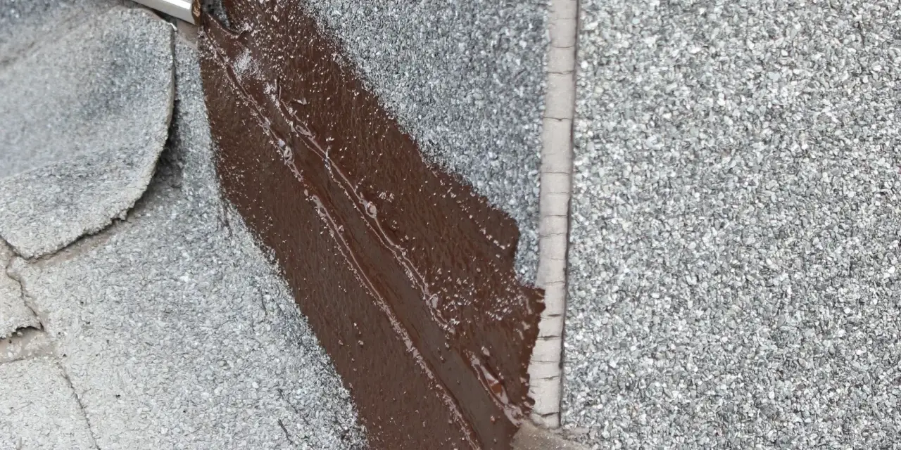 Fragment dachu pokryty papą z posypką, gdzie świeżo nałożona brązowa masa bitumiczna uszczelnia połączenie z metalowym elementem.