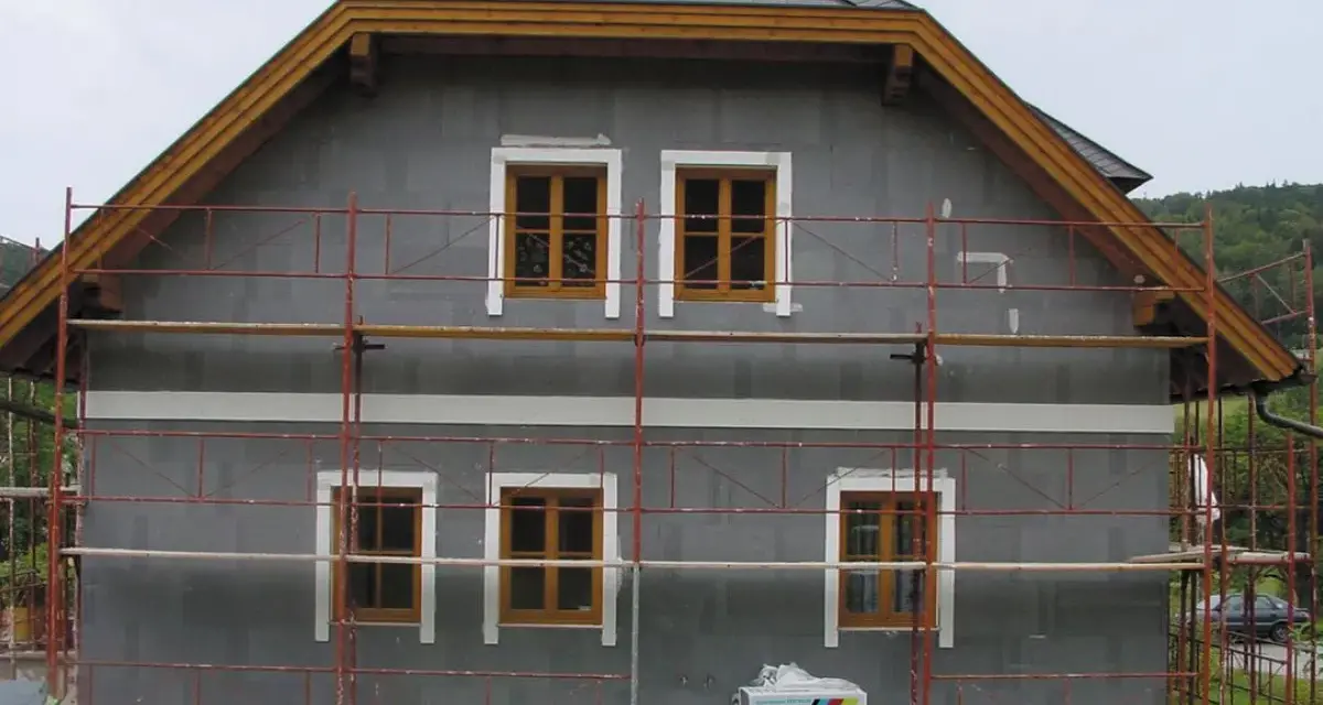 Dom w trakcie ocieplania. Szara elewacja, drewniane okna, rusztowanie. Materiały budowlane Austrotherm wskazują na dbałość o niski współczynnik przewodzenia ciepła.