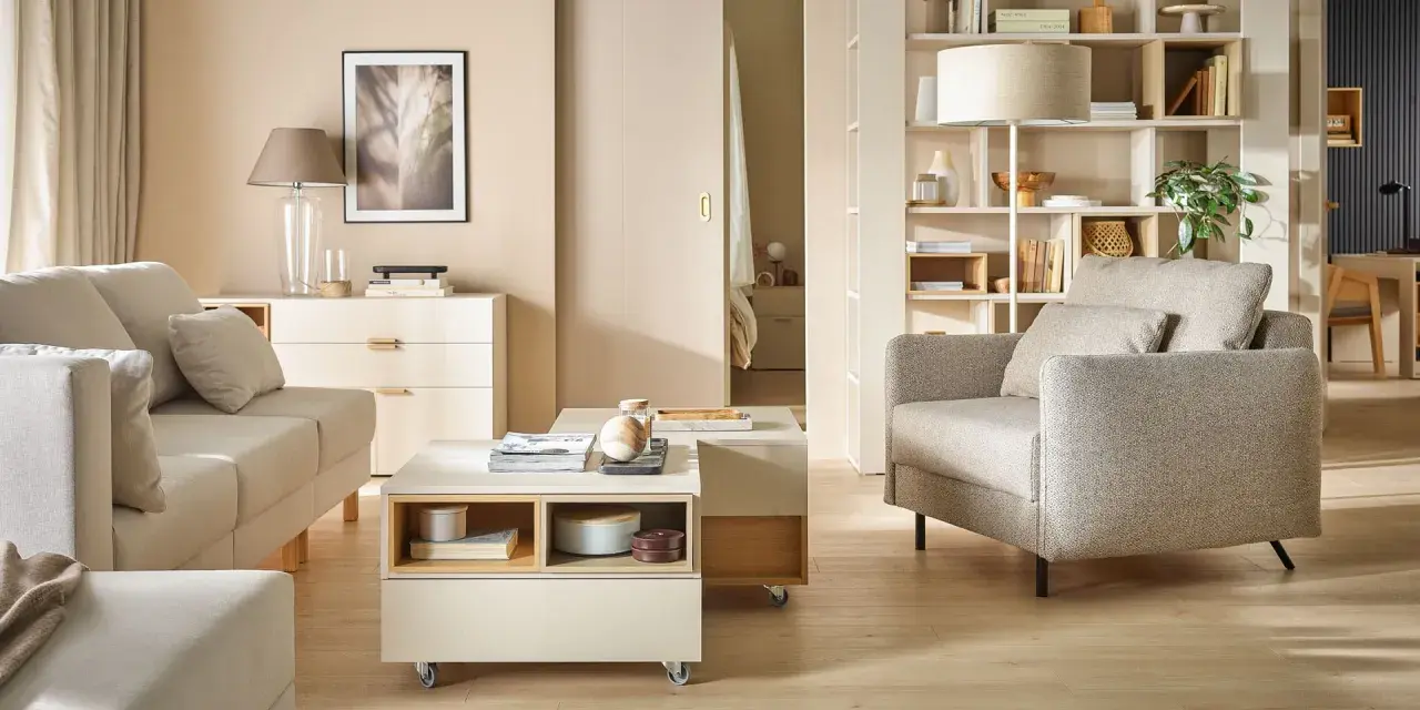 Jasny salon z sofą, fotelem i stolikiem kawowym na kółkach. Na półkach regału stoją dekoracje, książki i lampa. Wersaliki dodają elegancji.