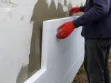 Pracownik w czerwonych rękawicach montuje styropian na ścianie. Dowiedz się, ile kosztuje elewacja domu.