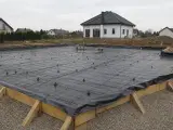 Fundament budowy domu z zbrojeniem i folią fundamentową, gotowy do zalania betonem. W tle widać domy mieszkalne.