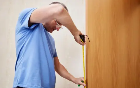 Mężczyzna w niebieskiej koszulce mierzy drzwi taśmą mierniczą.