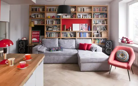Przytulny salon z regałem pełnym książek, szarą kanapą i czerwonym fotelem. Wnętrze nawiązuje do **stylu PRL** – czerwona lampa, plakaty i telefon dodają klimatu.