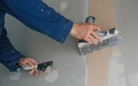 Ręce w niebieskim kombinezonie wykonują szpachlowanie ścian, wygładzając masę szpachlową za pomocą narzędzi.