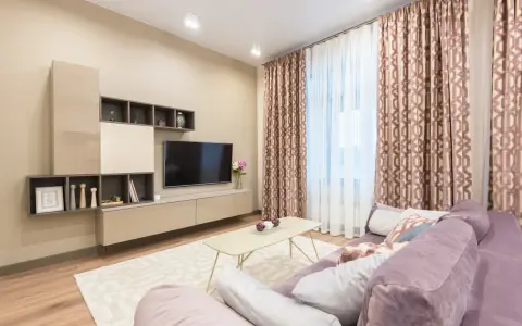 Przytulny salon z fioletową kanapą, beżowy kolor ścian tworzy ciepłą atmosferę. W tle telewizor i zasłony.