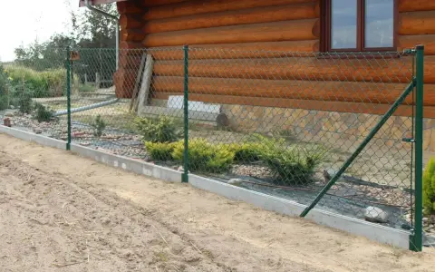 Montaż siatki ogrodzeniowej przy drewnianym domu. Widać zielone słupki, siatkę i betonowy podmurówkę.
