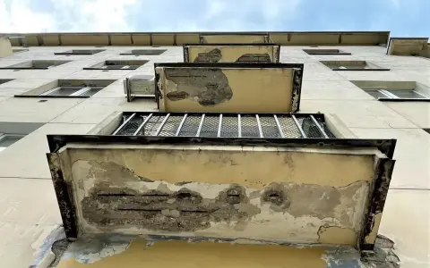 Zniszczone balkony bloku mieszkalnego, z odsłoniętym zbrojeniem, jakby czas był dla nich **inhibitorem** postępu.