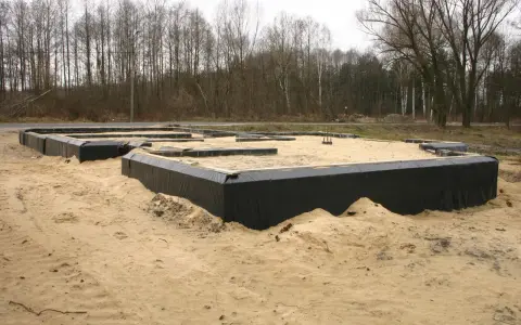 Fundament domu w budowie, otoczony czarną folią i piaskiem, na tle drzew.