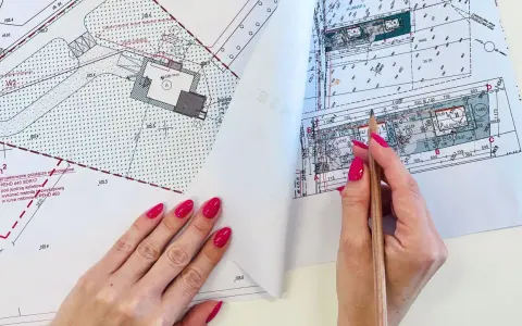 Dłoń z różowym lakierem na paznokciach trzymająca ołówek nad mapą projektową.