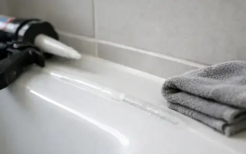 Pistolet do silikonu i świeża fuga na białej powierzchni. Zastanawiasz się, jak długo schnie silikon? Czas schnięcia zależy od wielu czynników.
