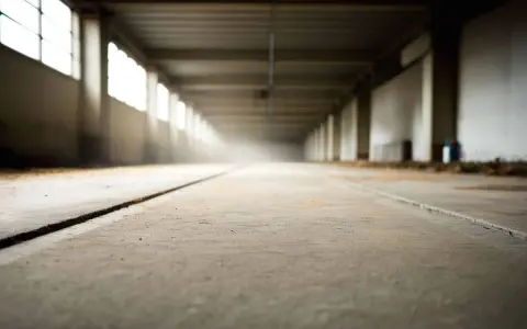Długi korytarz z betonową posadzką, gdzie trwa zacieranie betonu. Światło wpada przez okna, tworząc mglistą atmosferę.