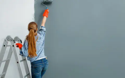Dziewczyna w kratkę i pomarańczowych rękawiczkach maluje ścianę wałkiem. Obok stoi drabina. Zastanawiasz się, ile farby na m2 zużyjesz?