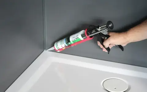 Dłoń trzyma pistolet z tubą **sylikonu**, aplikując go w rogu brodzika prysznicowego.