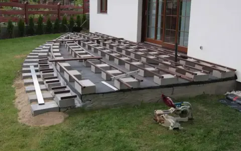 Budowa tarasu: betonowe bloczki i metalowe legary pod taras wymiary, gotowe do ułożenia desek. Obok piła.