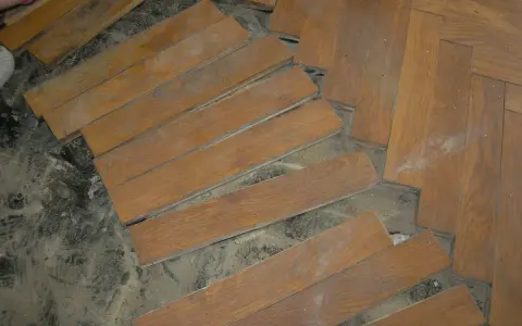 Drewniany parkiet w jodełkę, częściowo podniesiony, odsłaniający podłoże.