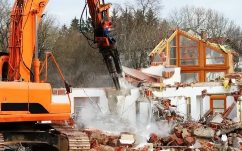 Pomarańczowa koparka niszczy dom, unosząc tumany kurzu i gruzu.
