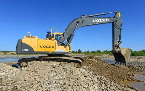 Żółta koparka Volvo EC240B LC pracuje na kamienistym brzegu rzeki. Zastanawiasz się, ile kosztuje godzina pracy koparki?