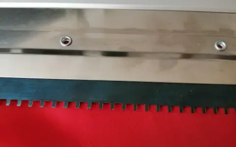 Metalowa rakla z ząbkowanym ostrzem, gotowa do pracy na czerwonym materiale.