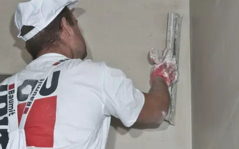 Murarz w białej czapce i koszulce z logo Baumit wygładza ścianę gładzią. Tynk jest nakładany kielnią.