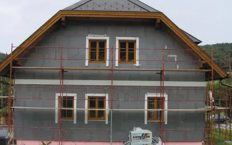 Dom w trakcie ocieplania. Szara elewacja, drewniane okna, rusztowanie. Materiały budowlane Austrotherm wskazują na dbałość o niski współczynnik przewodzenia ciepła.
