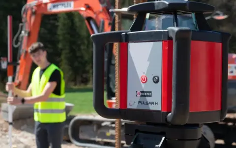 Niwelator laserowy Pulsar H na budowie. W tle pracownik w kamizelce odblaskowej i koparka.