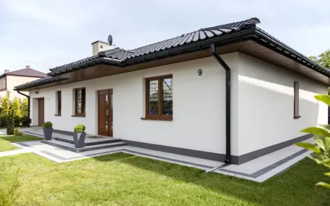 Nowoczesny biały dom z brązowymi oknami i drzwiami, otoczony zielonym trawnikiem.