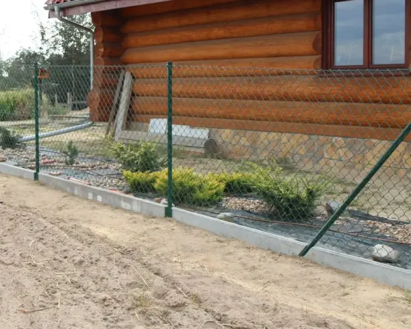 Montaż siatki ogrodzeniowej przy drewnianym domu. Widać zielone słupki, siatkę i betonowy podmurówkę.