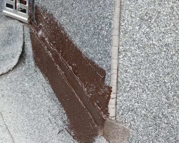 Fragment dachu pokryty papą z posypką, gdzie świeżo nałożona brązowa masa bitumiczna uszczelnia połączenie z metalowym elementem.
