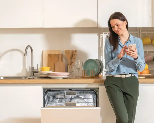 Kobieta sprawdza na telefonie, ile wody zużywa zmywarka, która jest otwarta i pełna naczyń.
