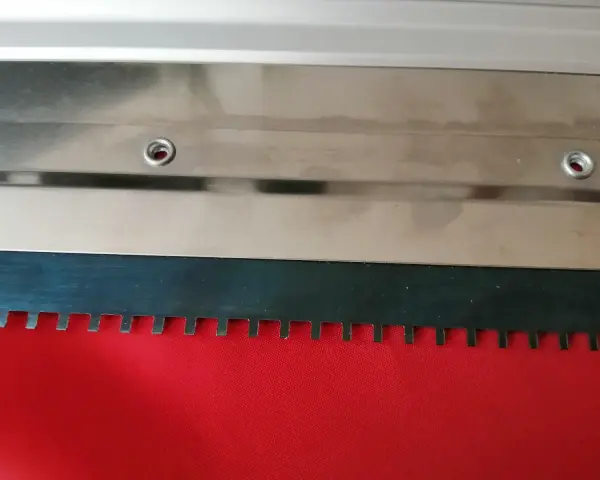 Metalowa rakla z ząbkowanym ostrzem, gotowa do pracy na czerwonym materiale.