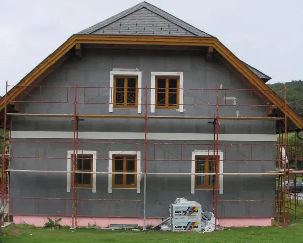 Dom w trakcie ocieplania. Szara elewacja, drewniane okna, rusztowanie. Materiały budowlane Austrotherm wskazują na dbałość o niski współczynnik przewodzenia ciepła.