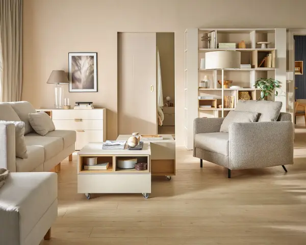 Jasny salon z sofą, fotelem i stolikiem kawowym na kółkach. Na półkach regału stoją dekoracje, książki i lampa. Wersaliki dodają elegancji.