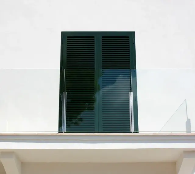 Nowoczesny balkon z przeszkloną balustradą i ciemnozielonymi okiennicami. Okapnik chroni przed deszczem.