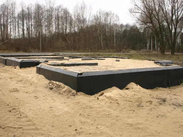 Fundament domu w budowie, otoczony czarną folią i piaskiem, na tle drzew.