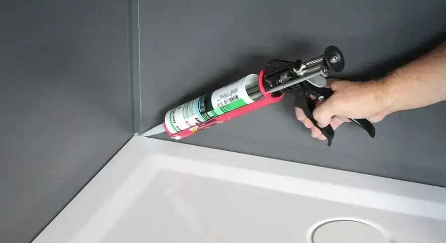 Dłoń trzyma pistolet z tubą **sylikonu**, aplikując go w rogu brodzika prysznicowego.