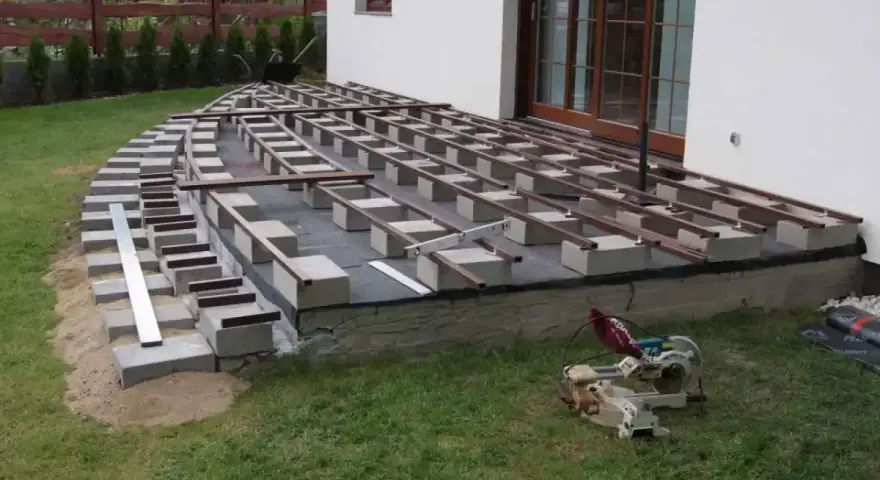 Budowa tarasu: betonowe bloczki i metalowe legary pod taras wymiary, gotowe do ułożenia desek. Obok piła.